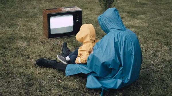 Yağmurluk giymiş küçük çocuklu bir kadın çimenlerde oturup retro televizyon izliyor.