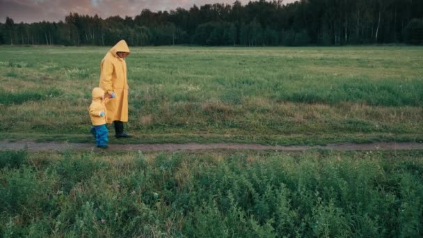 Wieczorem ojciec spaceruje po parku z małym dzieckiem. Ludzie w płaszczach przeciwdeszczowych — Wideo stockowe