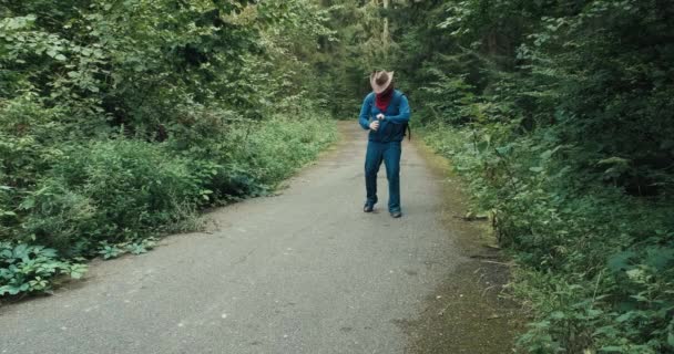 Dronken man met cowboyhoed loopt langs bosweg en drinkt alcohol uit een fles — Stockvideo