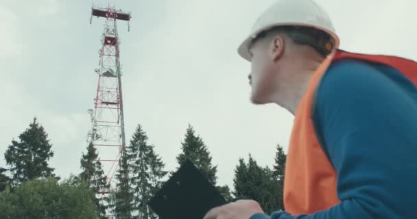 4G 5G κυψελοειδής πύργος ραδιόφωνο, εργαζόμενος σε φόρμες και κράνος στέκεται δίπλα του — Αρχείο Βίντεο