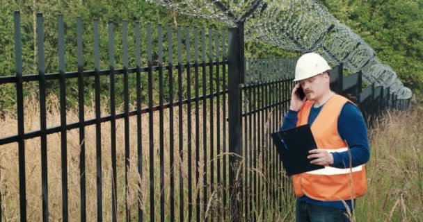 Builder is discussing something on phone standing next to fence with barbed wire — Stock Video