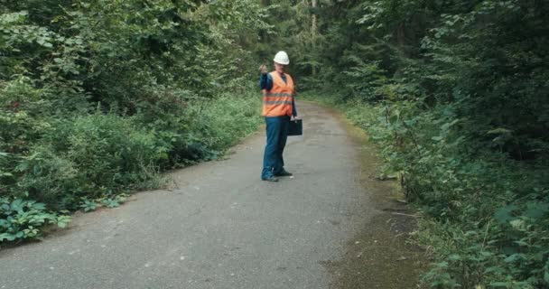 Trabajador en ropa de trabajo y un casco en una carretera en el bosque — Vídeos de Stock