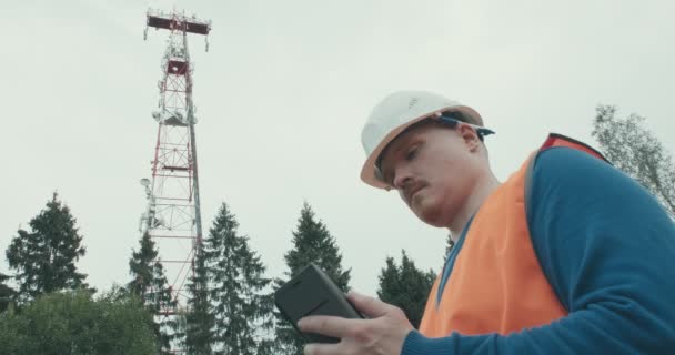 Obok stoi komórkowa wieża radiowa 4G 5G, pracownik w kombinezonie i hełmie — Wideo stockowe