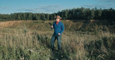 Kovboy şapkalı bir adam kumlu vadide dikiliyor ormanla kaplı. Sigara içiyor.