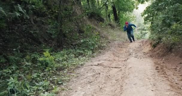Человек в красной бандане бежит крутой склон посреди летнего леса — стоковое видео