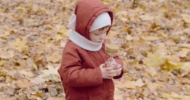 Kleines kaukasisches Mädchen spielt im herbstlichen Wald mit einer gläsernen Sanduhr — Stockvideo