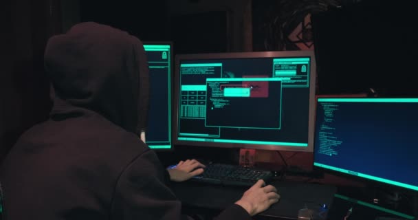 Hacker in a hood sits in front of computer screens and hacks databases — Vídeo de Stock