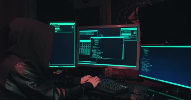 Hacker in a hood sits in front of computer screens and hacks databases — Vídeo de Stock