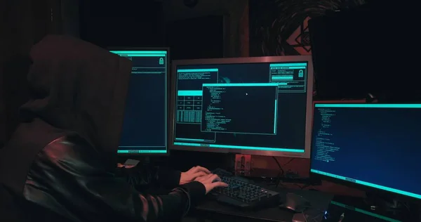 Hacker in a hood sits in front of computer screens and hacks databases — Stock Photo, Image