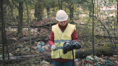 İş kıyafetleri ve kask giyen çevre bilimci kadın ormandaki çöpleri temizliyor.