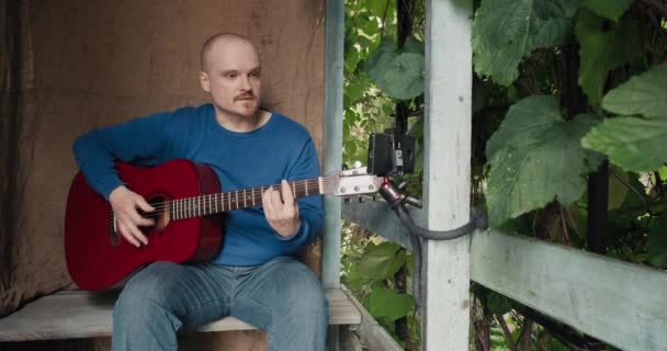 Mannen med gitarr sitter på verandan och sjunger låtar medan han läser text från sin telefon — Stockvideo