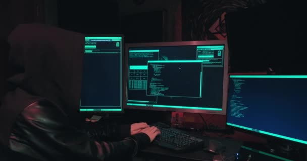 Hacker σε μια κουκούλα κάθεται μπροστά από οθόνες υπολογιστών και αμυχές βάσεις δεδομένων — Αρχείο Βίντεο