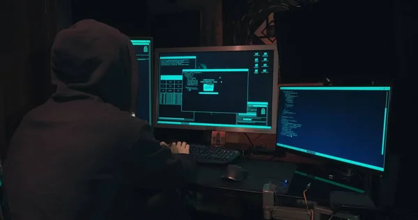 Hacker en una capucha se sienta frente a las pantallas de la computadora y hacks bases de datos —  Fotos de Stock