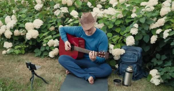 Человек путешествует с гитарой, сидит в парке в природе и проводит онлайн-трансляцию — стоковое видео