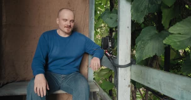 Człowiek komunikuje się online za pomocą telefonu, siedząc na ganku wiejskiego domu — Wideo stockowe