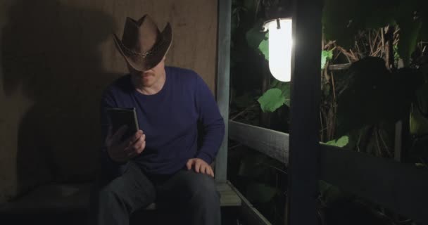 Cowboy man communiceert online met zijn telefoon terwijl hij op de veranda zit. Welterusten — Stockvideo