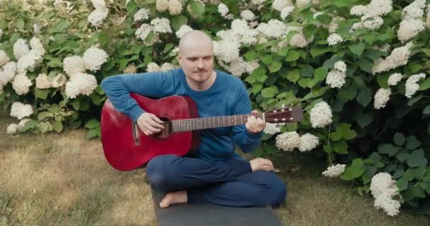 Vit man sitter i en park bland blommor och spelar röd akustisk gitarr — Stockvideo