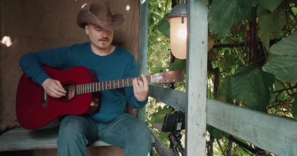 Человек с акустической гитарой сидит на крыльце деревенского дома и ведет онлайн подкаст — стоковое видео