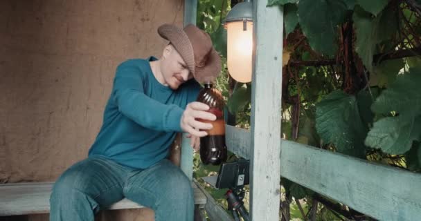 Ковбой на крыльце пьет пиво из бутылки и общается онлайн по телефону — стоковое видео