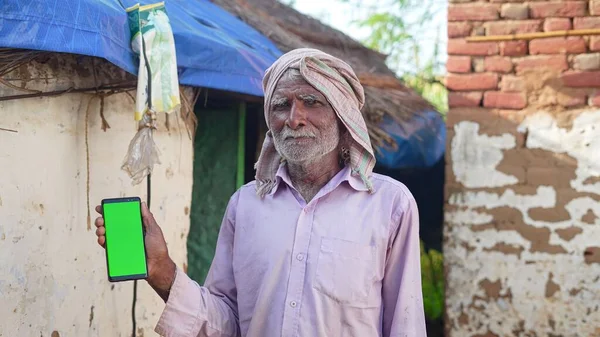 Indian old man holding a mobile phone and point towards it's screen. Mobile phone with a green screen for mockup.
