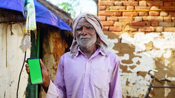 Indian old man holding a mobile phone and point towards it\'s screen. Mobile phone with a green screen for mockup.
