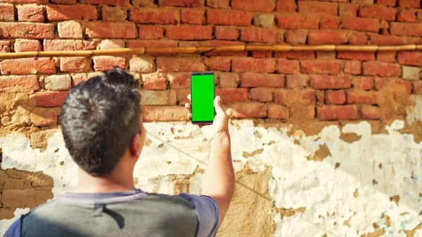 Young Indian man taking selfie and showing a green screen cell phone with rural village life concept background.