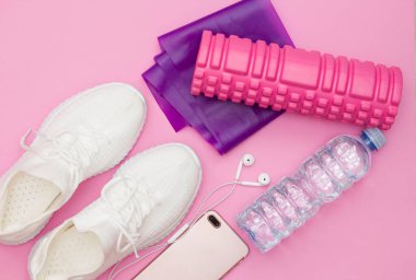 Su şişeli bir fitness konsepti, spor ayakkabıları, kulaklıklı bir telefon, mor bir fitness lastiği, pembe arka planda pembe bir silindir. Boşluğu kopyala.