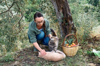 Bahçesindeki zeytin ağacının yanında bir sepet dolusu olgun hurma taşıyan lastik çizmeli bir kadın kedileriyle oynuyor. Bahçede çalışmak yeni normalin hobisi. Hasat et. Düş! Sinerjik bahçe.