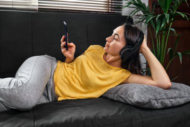 Kablosuz kulaklıkla müzik dinleyen ve kanepede uzanmış akıllı telefon kullanan bir kadın..