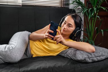 Kablosuz kulaklıkla müzik dinleyen ve kanepede uzanmış akıllı telefon kullanan bir kadın..