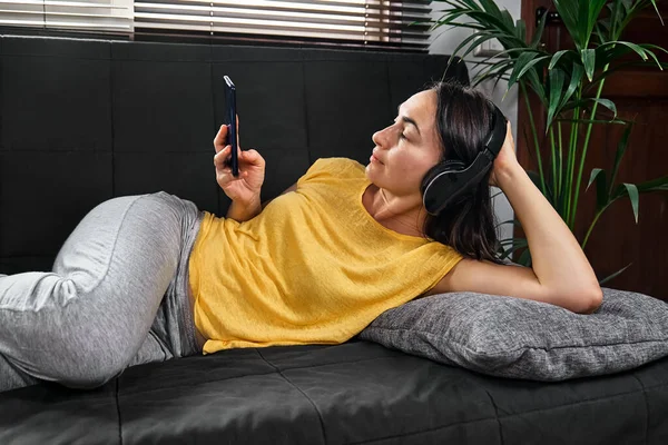 Kablosuz kulaklıkla müzik dinleyen ve kanepede uzanmış akıllı telefon kullanan bir kadın..