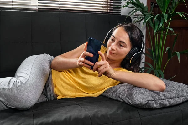Kablosuz kulaklıkla müzik dinleyen ve kanepede uzanmış akıllı telefon kullanan bir kadın..