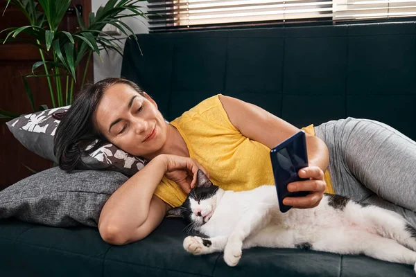 Brunette woman lying with her cat on the sofa uses the mobile phone. People with gadget. Communication mobile.