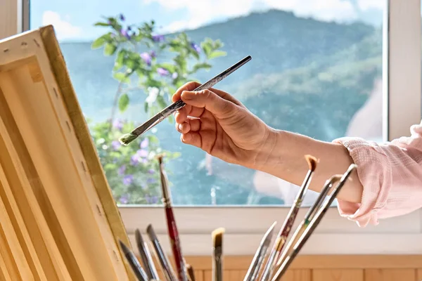 Female hand holding a paintbrush near canvas on easel near the window in the studio. Art painting hobby. Creative leisure. Self expression concept.