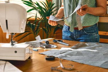 Kadın keten kumaş desenli bir kağıt kesiyor. Küçük stüdyodaki dikiş makinesinin terzisi. Moda atölyesi, terzilik, el yapımı kıyafet konsepti. Yavaş Moda Bilinci Tüketimi