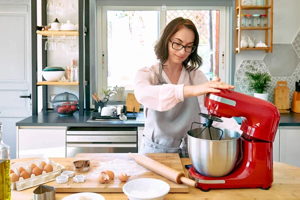 Sıcak bir mutfakta önlük giymiş kurabiye pişiren bir kadın. Ev hanımı, gezegen mikserindeki kekin hamurunu yapıyor. Ev yapımı kekler veya turtalar..