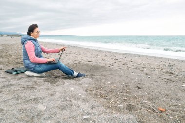 Diz üstü bilgisayarlı gözlük takan güzel, sportif bir kadın deniz kenarında kış kumsalında otururken dışarıda çalışıyor. Modern yaşam tarzı, bağlantı, blog yazma, iş dünyası, serbest çalışma.