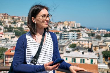İtalya, Sicilya 'daki Messina manzarasının tadını çıkarırken cep telefonunda müzik, şarkı ya da ses rehberi dinleyen mutlu bir kadın. Kentsel seyahat eğlencesi.