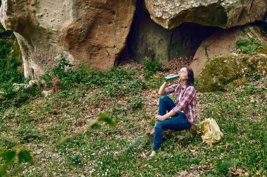 Ormanda dinlenmek için bir taşın üzerinde oturan bir kadın, sırt çantasıyla bir korulukta seyahat ediyor ya da dışarıda egzersiz yapıyor. Aktif insanlar, yürüyüş, sağlıklı yaşam tarzı ve uyum kavramı. Doğayla birlik.