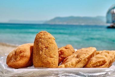 Typical Sicilian street food on seaside background. Hot palatable panzerotti (calzone pizza fried in hot oil) and arancina (deep fried rice balls with meat) on paper plate take out.