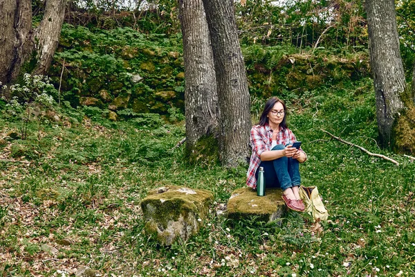 Ormanda bir taşın üzerinde oturan bir kadın, konuşmak için cep telefonu kullanıyor ya da GPS arıyor. Sırt çantasıyla seyahat etmek ya da dışarıda egzersiz yapmak. Aktif insanlar, yürüyüş, sağlıklı yaşam tarzı ve uyum kavramı.