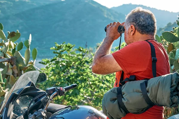 On the road. Tourist man traveling on motorcycle, stopped for looking through binoculars at the mountains view. Active people, hiking, healthy lifestyle and harmony concept. Unity with nature.