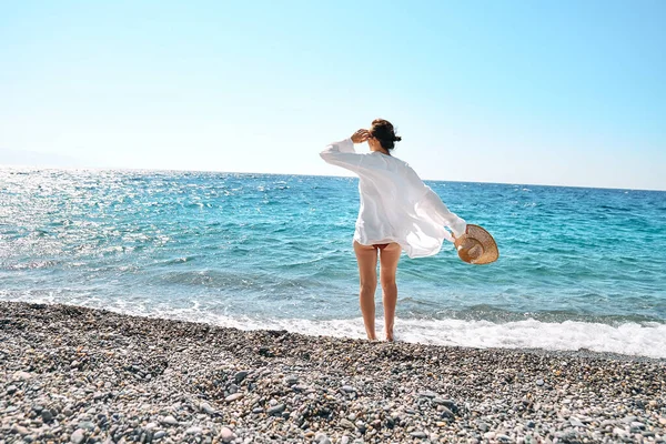 Hasır şapkalı, beyaz gömlekli, çakıl taşlı plajda yürüyen kaygısız bir kadın. Yazın deniz kenarında dinlenip tatilin tadını çıkarıyorum. Doğayla birlik. Sağlık, başarı, özgürlük ve seyahat kavramı