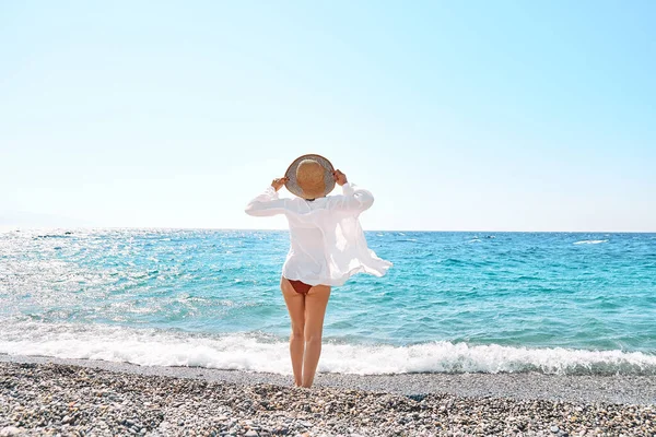 Back view of carefree woman in straw hat and white shirt walking on pebble beach. Relaxing and enjoy holiday at the sea in summertime. Unity with nature. Wellness, success, freedom and travel concept