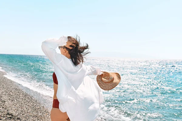 Carefree woman in white shirt and straw hat standing in beach in windy day. Relaxing and enjoy holiday at the sea in summertime. Unity with nature. Wellness, success, freedom and travel concept.