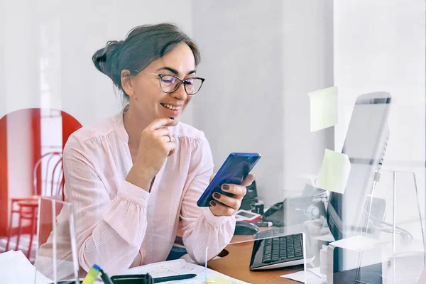 Smiling middle-aged business woman has video chat on mobile phone, sitting in office at desk with glass partition.Female manager worker use smartphone online apps for business social media management.