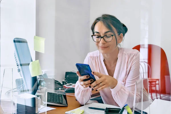 Smiling businesswoman received good news on mobile phone, sitting in office at desk with glass partition.Female manager worker use smartphone online apps for business social media management.