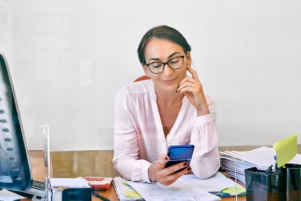 Middle-aged businesswoman checking email messages on mobile phone sitting in office at desk with glass partition.Female manager worker use smartphone online apps for business social media management