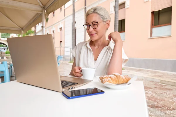 Female freelancer working on laptop at cafe. Portrait of young blond smiling woman sitting alone in cafe outdoors and has video conference in her notebook computer.