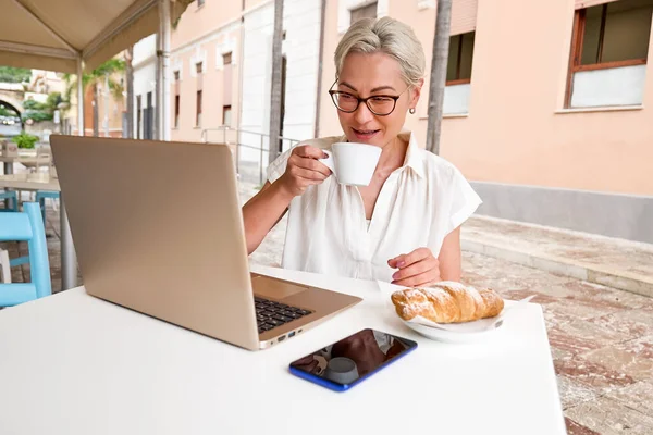 Female freelancer working on laptop at cafe. Portrait of young blond smiling woman sitting alone in cafe outdoors and drinking coffee while has video conference in her notebook computer.